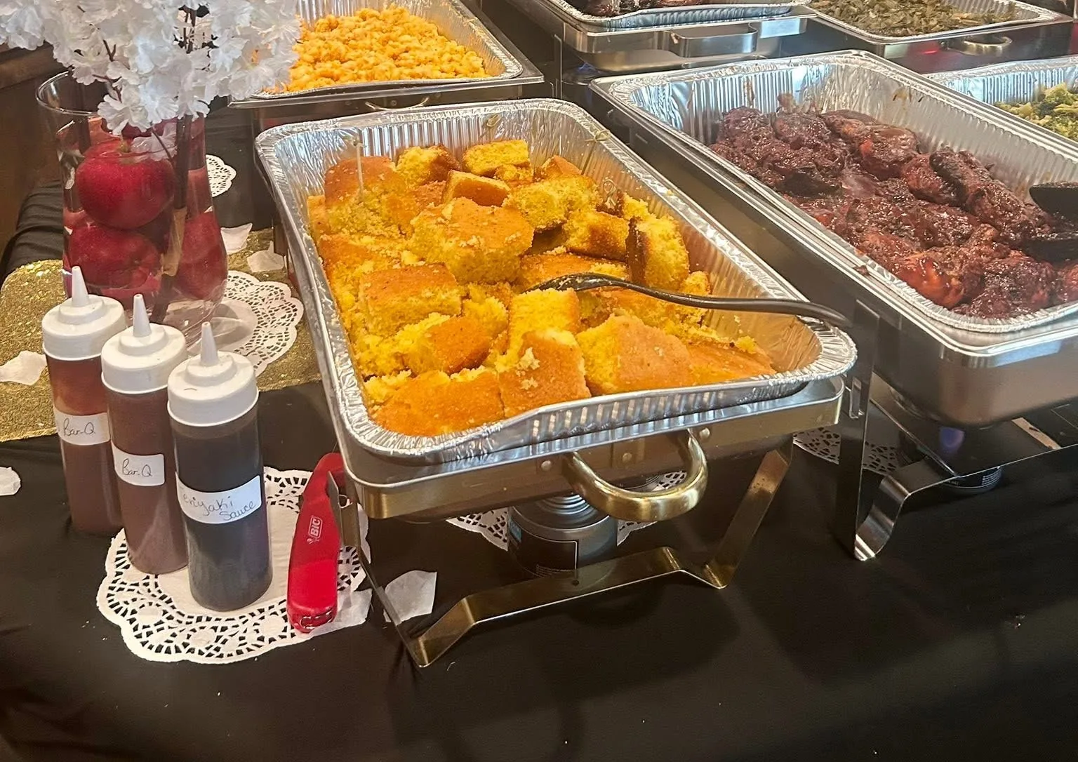 A tray of butter cornbread beside barbecue chicken and sauces on a draped catering table