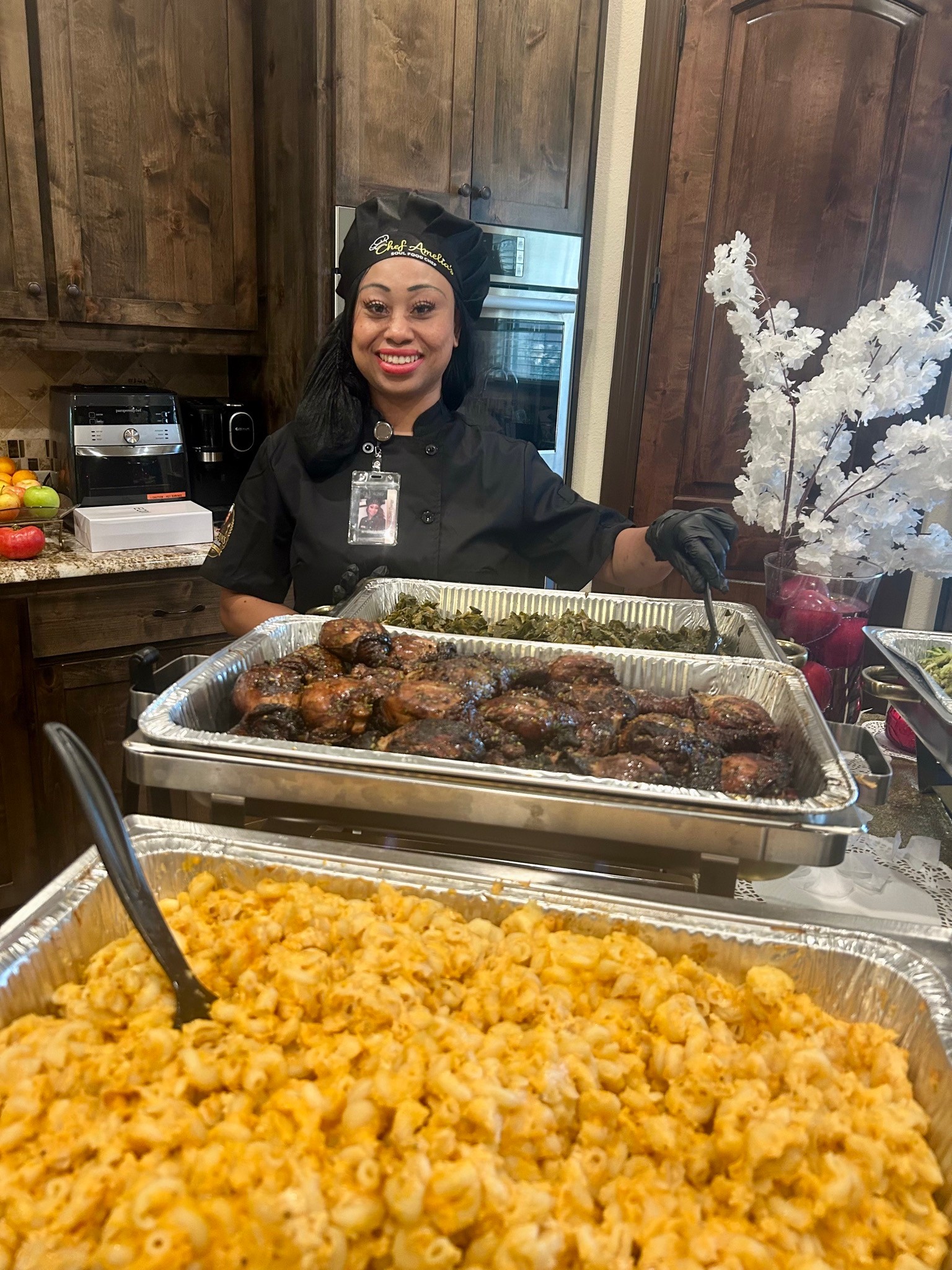 Chef Amelia in a black chef’s coat stands behind trays of macaroni and cheese, glazed chicken, and greens.