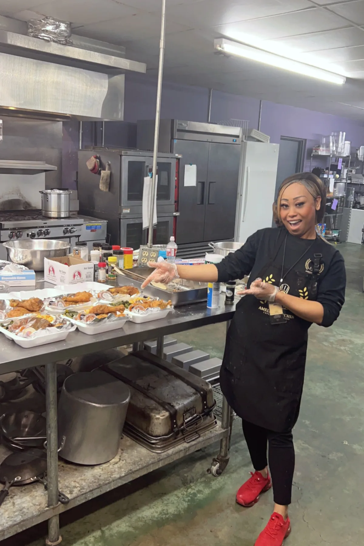 Chef Amelia laughing as she gestures toward a row of packed plates in her commercial kitchen